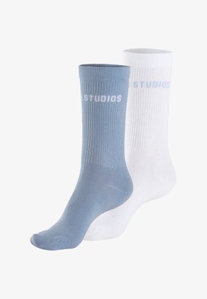 Bombažne kratke nogavice v svetlo modri in beli barvi. Žlebasta tekstura z napisom 'STUDIOS' v belem na modri nogavici, ravna zasnova prstov.