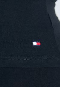 Zwarte katoenen shirt met een klein geborduurd logo in rood, wit en blauw op de linkerbovenhoek, met een ribbeltextuur en zoom.