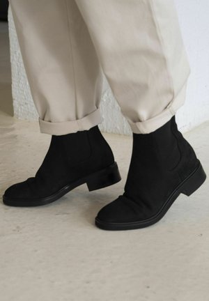 Stivali ankle in suede nero con una finitura liscia, punta arrotondata e tacco basso a stack, abbinati a pantaloni beige risvoltati su un pavimento neutro.