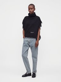 Personne debout portant un poncho noir à col roulé avec des franges, un jean bleu clair coupe décontractée et des mocassins noirs brillants sur un fond uni.