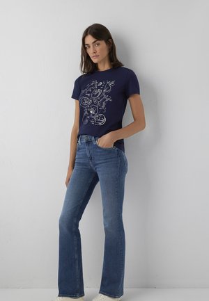 Young woman with dark hair wearing a navy blue t-shirt with white cat illustrations and blue jeans, standing against a white wall.
