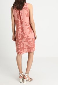 Rosa ärmelloses Kleid mit floralem Muster, gefertigt aus glattem Stoff. Kombiniert mit weißen Sandalen mit Absätzen, die ein schlichtes Design aufweisen.