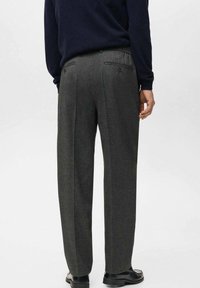 Pantalones gris claro a medida con un acabado texturizado, un bolsillo trasero único y doble pinzas, combinados con un suéter oscuro, vistos desde la espalda.