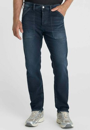 Man wearing dark blue straight-leg jeans with large front pockets and silver-gray athletic shoes, standing with one hand in pocket.