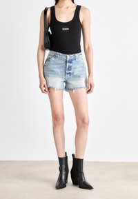 Kvinna iklädd svart linne, ljusblå denimshorts, svarta ankelskor och bärande liten svart axelväska, stående på grått golv.