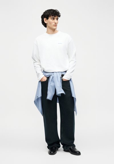 Νέος άνδρας με σγουρά μαλλιά που φοράει λευκό φούτερ, σκουρόχρωμο παντελόνι με φαρδιά μπατζάκια, μαύρα παπούτσια και μπλε ριγέ πουκάμισο δεμένο γύρω από τη μέση του.