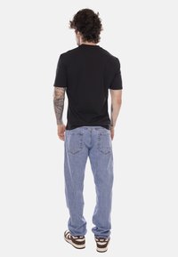 Uomo con tatuaggi che indossa una maglietta nera, jeans azzurri chiari e sneakers Nike marroni, in piedi di spalle su uno sfondo bianco.