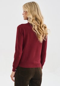 Frau mit langen, welligen blonden Haaren, die einen weinroten Langarmpullover und dunkelbraune Hosen trägt, seitlich vor einem schlichten Hintergrund stehend.