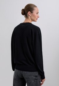 Vrouw met een messie in een knot draagt een zwarte trui met een rafelige kraag en donkergrijze jeans, zijwaarts gekeerd tegen een effen grijze achtergrond.