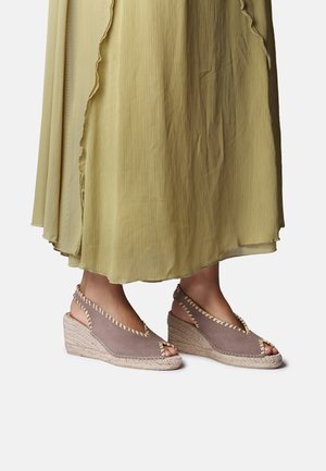 Fødder iført taupe kilehæl espadrille sandaler med beige syning kombineret med en flydende lys grøn plisseret nederdel.