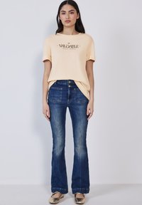 Camiseta amarillo claro con texto negro "VALUABLE", combinada con jeans acampanados de color azul oscuro, que cuentan con bolsillos frontales y una cintura alta. Bailarinas doradas.