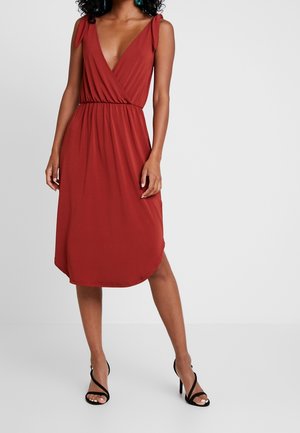 Femme portant une robe sans manches, rouge rouille, longueur genou, avec un décolleté en V profond et une taille froncée, assortie à des sandales noires à talons hauts.