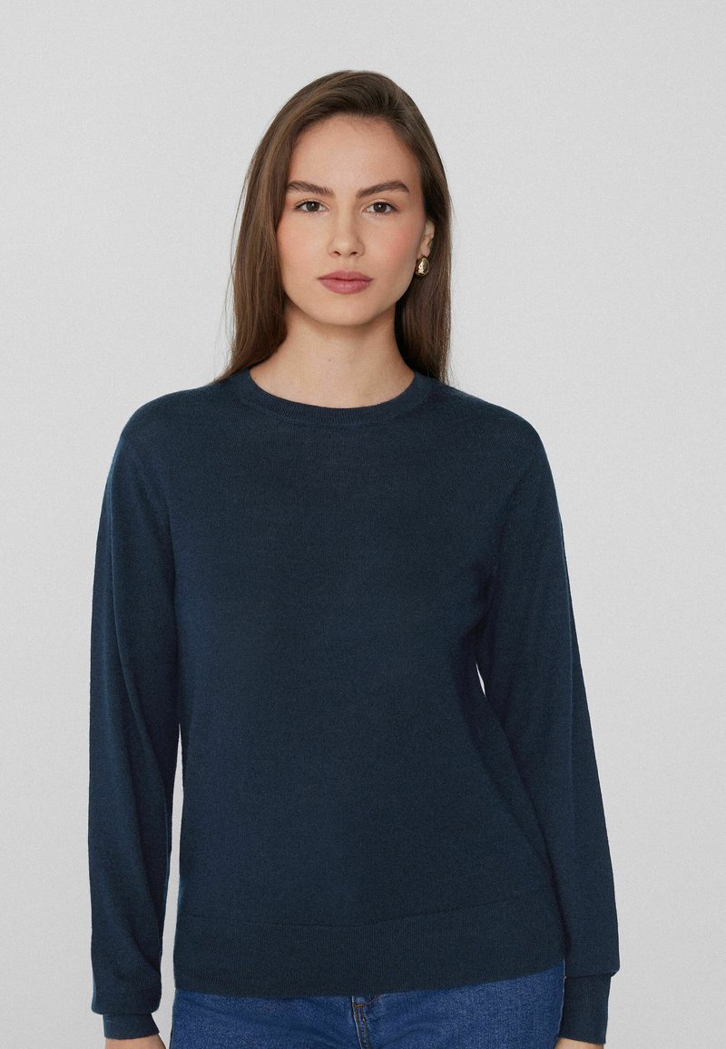 Suéter de punto azul marino con mangas largas, cuello redondo y textura suave. Ajustado en el dobladillo, combinado con jeans de mezclilla.