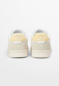 Sneaker bianche e beige con un accento in suede giallo sul tallone, dettaglio del logo circolare e suola testurizzata con cuciture visibili.