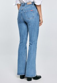 Žena nosící světle modré rozšířené džíny s zadními kapsami a černé kotníkové boty na podpatku, stojící před jednoduchým pozadím.