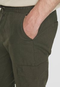 Dunkelgrüne Hosen aus leichtem Stoff, mit einem elastischen Bund und Seitentaschen, sowie einer Cargo-Tasche am Oberschenkel.