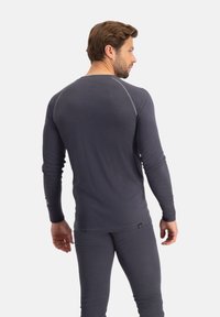 Man die wegkijkt en een nauwsluitend donkergrijs shirt met lange mouwen en een broek draagt tegen een effen lichte achtergrond.