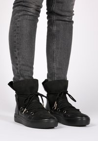 Zapatillas negras de caña alta con parte superior de ante, suela de goma, forro de pelo en el puño y detalles de cordones con adornos plateados.