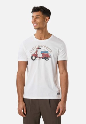 Ung mand smilende, iført en hvid t-shirt med en rød vintage scooter grafik og beige bukser, stående mod en ensfarvet baggrund.