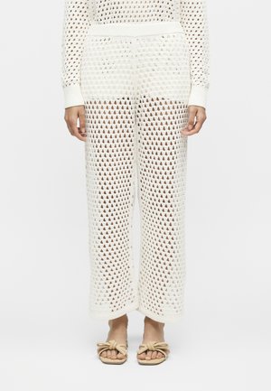 Osoba nosząca białe szydełkowane spodnie z szerokimi nogawkami oraz pasujące sandały z kokardkami, stojąca na jednolitym białym tle.