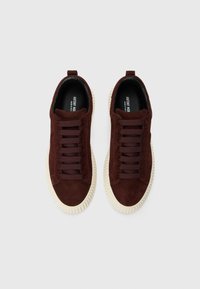 Une paire de baskets en daim marron foncé avec de grosses semelles en caoutchouc blanc et des lacets marron assortis, vue de dessus sur un fond uni.