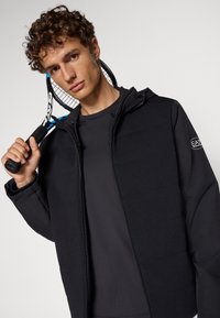 Haut zippé noir avec un tissu texturé, capuche et patch logo. Le modèle tient une raquette de tennis, portant un t-shirt sombre en dessous.
