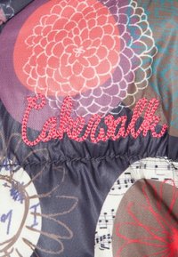 Kleurrijke stof met bloemige patronen in roze en grijs, met roze geborduurde tekst "Cakewalk" aan de naad.