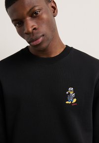 Hanorac negru cu un personaj de desene animate brodat, îmbrăcat într-o uniformă de pilot. Gulerul și manșetele răsucite oferă textură și structura.