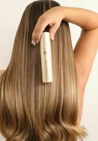 En guld sprayflaska hålls mot långt, rakt, ljusbrunt hår med subtila höjdpunkter, vilket visar en jämn textur och en glänsande yta.