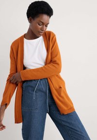 Orange knit cardigan with open front, ribbed cuffs, and two front pockets, worn over a white t-shirt and blue denim pants.