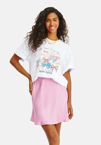 Biely grafický tričko s farebnými potlačami mašlí a textom, spárované s svetloružovou sukňou v A-črevníka, tvoriace neformálny outfit.