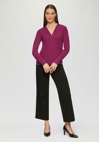 Top avvolgente aderente a maniche lunghe di un vivace colore viola, abbinato a pantaloni neri a vita alta e a gamba larga, e a tacchi alti neri. Tessuto liscio, design minimalista.