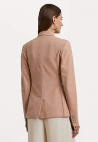 Blazer ajusté rose clair en tissu texturé, présentant une silhouette cintrée, des revers en pointe et des boutons décoratifs sur les manches.