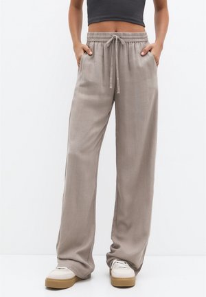 Persoon draagt losse beige broek met trekkoord en zijzakken, witte sneakers, cropped zwart top zichtbaar vanaf de taille naar beneden.