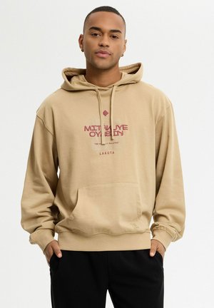 Joven con capucha beige con bolsillo delantero y texto "MIDWAY OYSTIN LAKOTA" de pie con las manos en los bolsillos sobre fondo blanco.