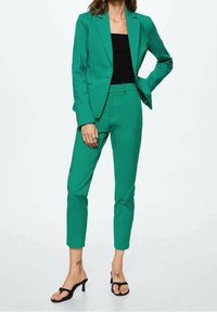 Costume vert ajusté avec une veste cintrée et un pantalon slim. Le matériau est lisse et présente un design épuré. Porté avec un haut noir et des sandales à talons.