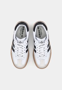 Un par de zapatillas Adidas blancas con rayas negras y suelas de goma, vistas desde arriba sobre un fondo blanco liso.