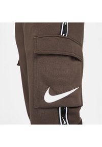 Bruna joggers med en cargofick, som har en framträdande vit Nike-logotyp och svarta sidostråkar med ett upprepat Nike-mönster.