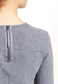 Grå texturerad topp med dragkedja bak, slät flik, rundad halsringning och subtila sömdetaljer.