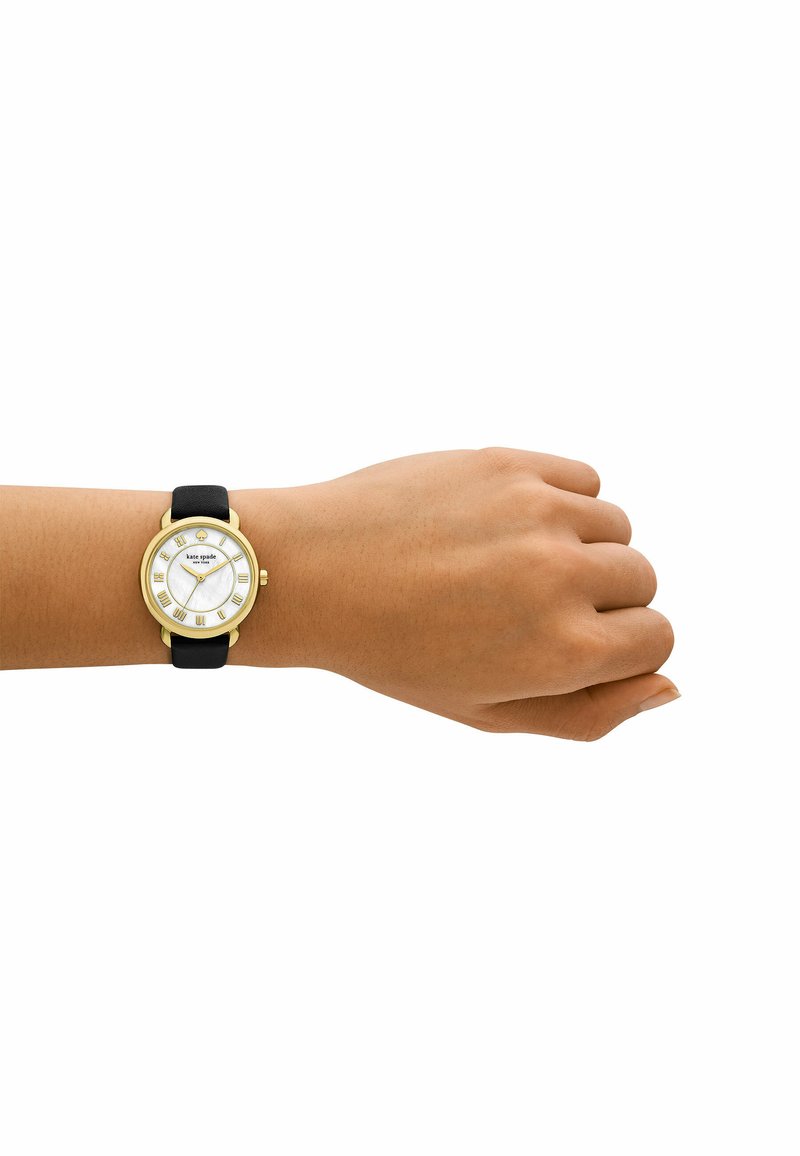 Guldtonad klocka med en vit snäcka urtavla, svart läderrem och romerska siffror, visad på en hand.