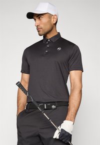 Čierne polo tričko s krátkymi rukávmi, gombíkmi a logom; v kombinácii s čiernym opaskom a šedými nohavicami; drží golfovú palicu; má na sebe bielu čiapku.