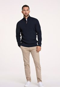 Marineblauwe kabeltrui met een kwart-rits kraag, gecombineerd met beige broeken en witte sneakers. De trui heeft ribgebreide manchetten en een ribgebreide zoom.