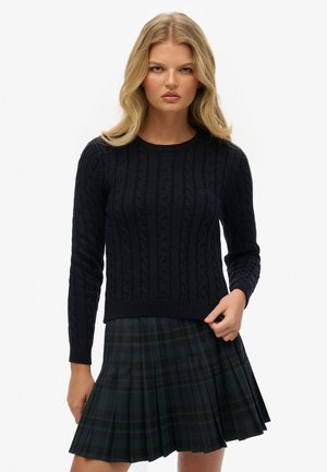 Marineblå kabelstrikket sweater med lange ærmer og ribbet kant, parret med en plisseret mørkegrønternet nederdel med sorte detaljer.