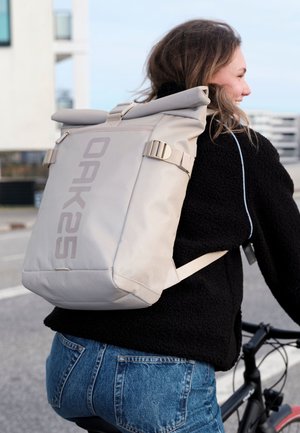 Femeie mergând pe bicicletă cu un rucsac bej cu închidere rulabilă cu marca „OAK25” și o jachetă polar neagră pe o stradă urbană.