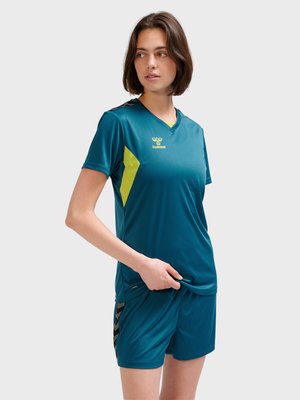 Kobieta w morsko-żółtym stroju sportowym stoi z ręką chwytającą koszulkę, patrząc w bok na jednolitym tle.