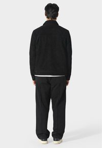 Černá semišová bunda s límcem, s detailem švu na zadní straně, nošena přes černé kalhoty. Model stojí zády, ukazuje hladkou texturu a minimalistický design.