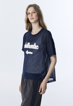 Jonge vrouw die een doorschijnend marineblauw shirt draagt met de tekst "Authentic" over een donker topje, gecombineerd met losse donkergrijze broek met zijstrepen.