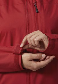 Rode jas met rits aan de voorkant. Close-up van handen die de klittenbandsluiting op de mouw aanpassen. Soepele stof, slim fit ontwerp en gestroomlijnde uitstraling.