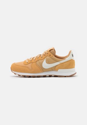Buty sportowe Nike w kolorze beżowym i jasnobrązowym z białym logo Swoosh, chropowatą cholewką z zamszu i tkaniny oraz żłobioną gumową podeszwą, widoczne z boku.