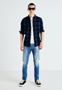 Îmbrăcăminte casual formată dintr-o cămașă flanel cu carouri albastre și negre, un tricou alb, blugi ușor albaștri și adidași negri cu tălpi albe. Ochelari de soare purtați.